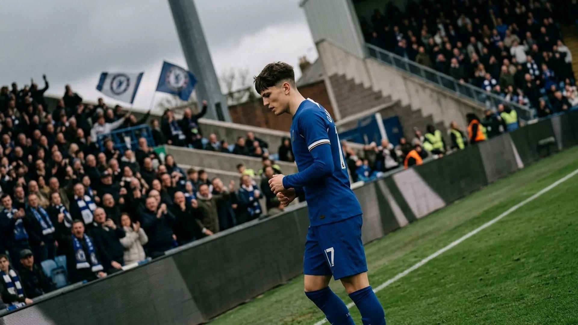 A football player in a Chelsea kit stands on the pitch, looking thoughtful, with blurred fans in the background.