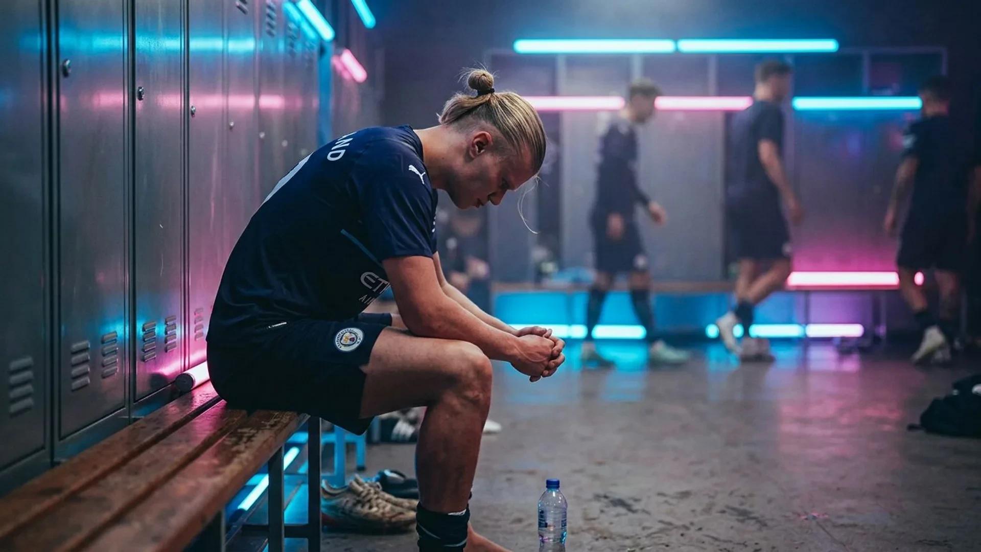 En nedslått Erling Haaland sitter alene i en mørk garderobe opplyst av neonlys, med tankene sine.