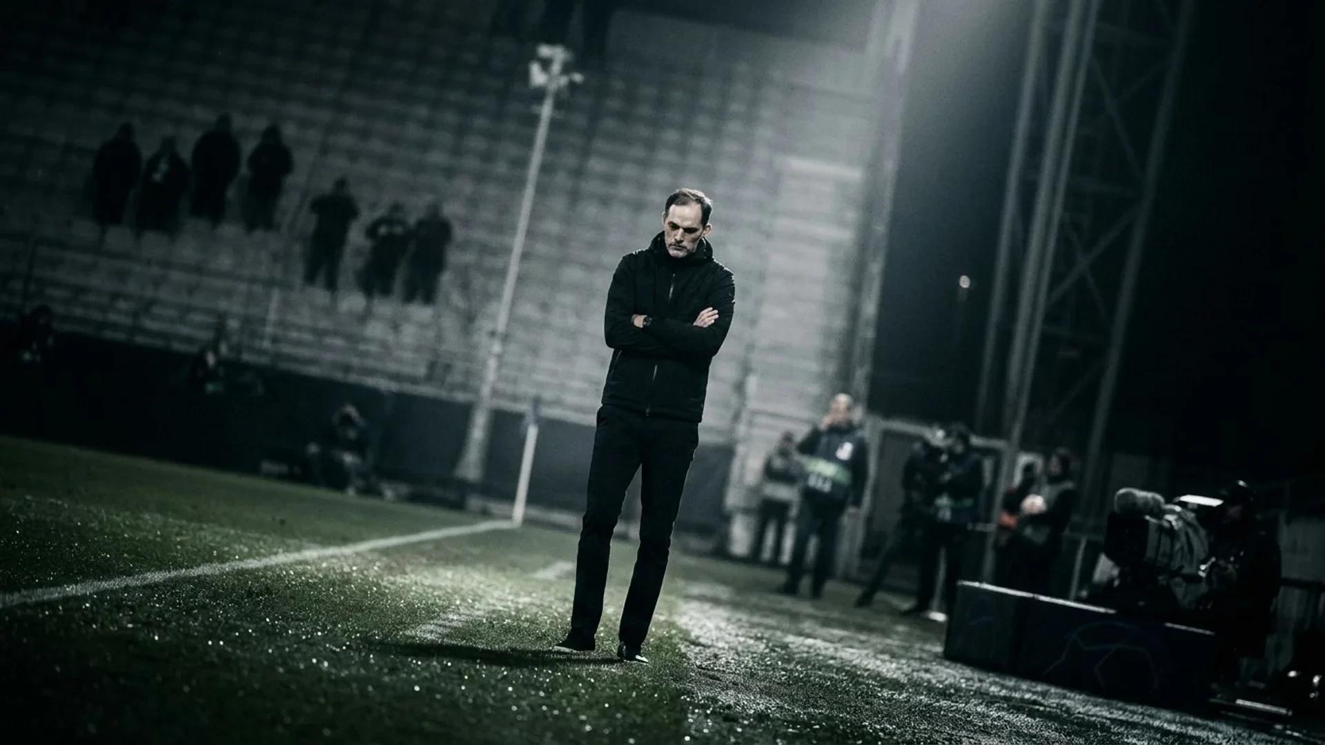 A somber Thomas Tuchel stands on the pitch in shadow, illuminated by a stark spotlight, reflecting on defeat.