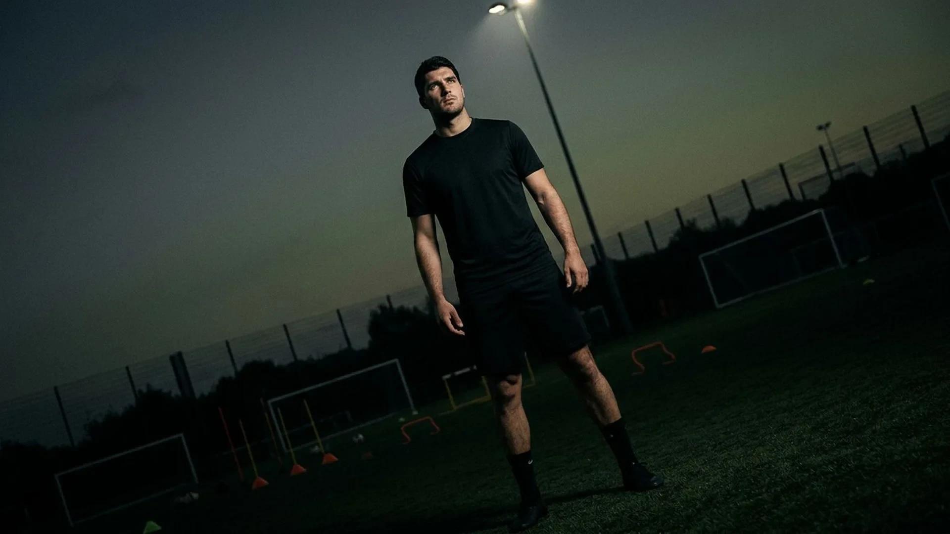 A tall football defender standing confidently in a training ground with dramatic lighting.