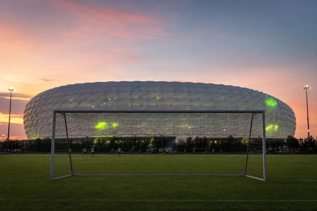 A wide shot of the Allianz Arena lit at dusk, with a goal post in the foreground and a pastel sky above.