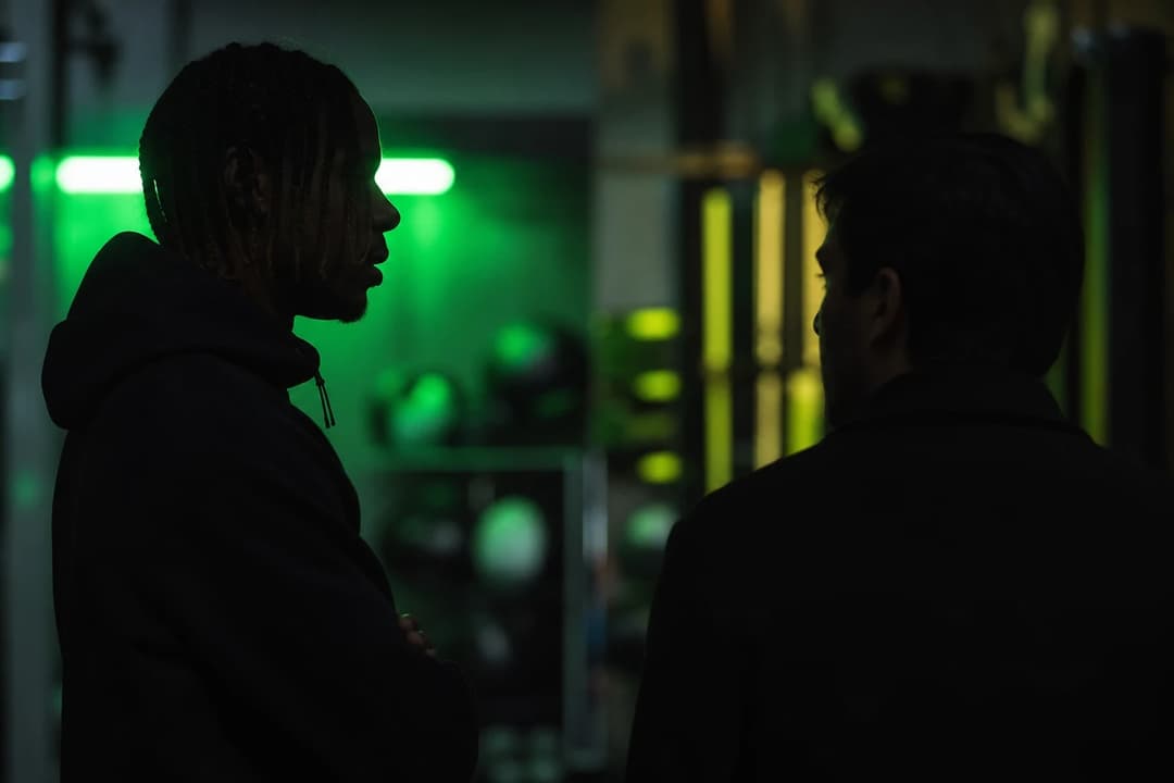 A shadowy figure, representing Bradley Barcola, speaks with a manager in a softly lit training facility with neon accents.