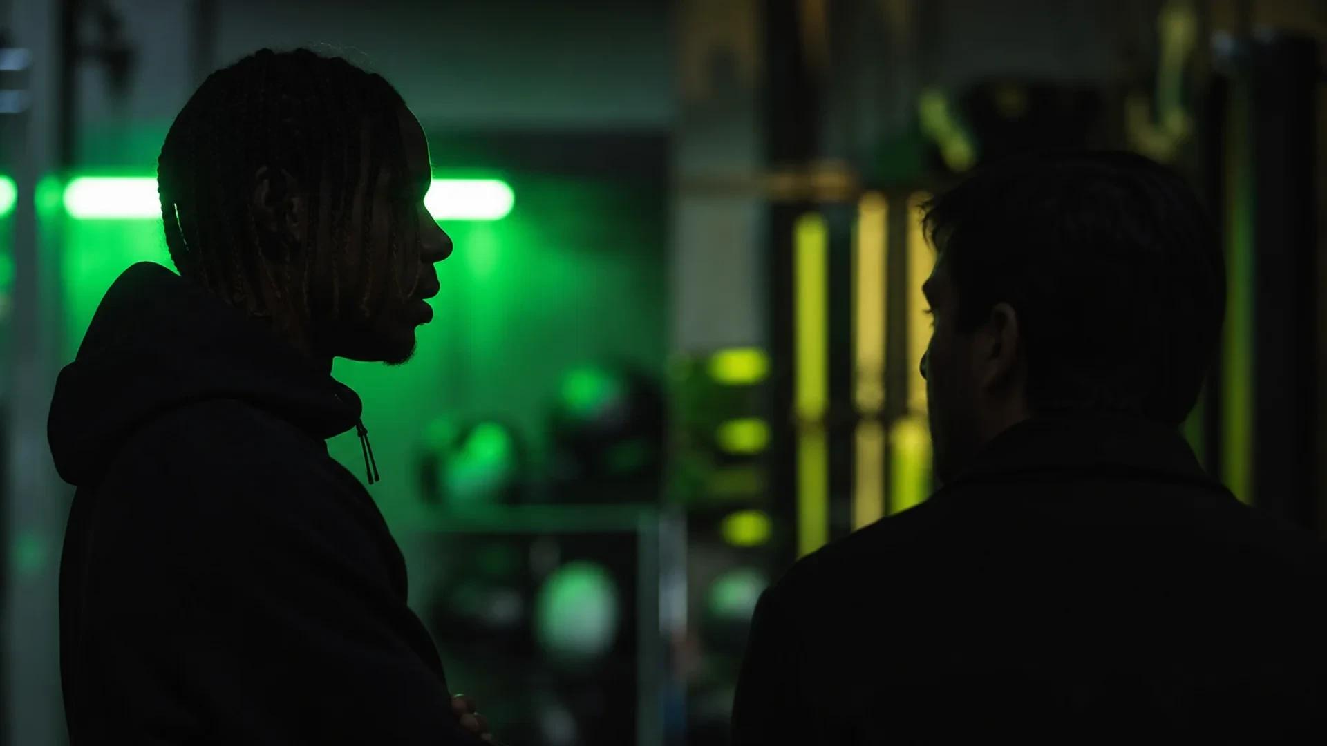 A shadowy figure, representing Bradley Barcola, speaks with a manager in a softly lit training facility with neon accents.