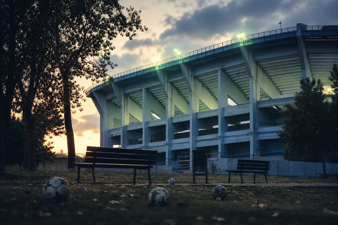 Wide shot of the empty Vallecas stadium with footballs in the foreground, sunlight filtering through trees.