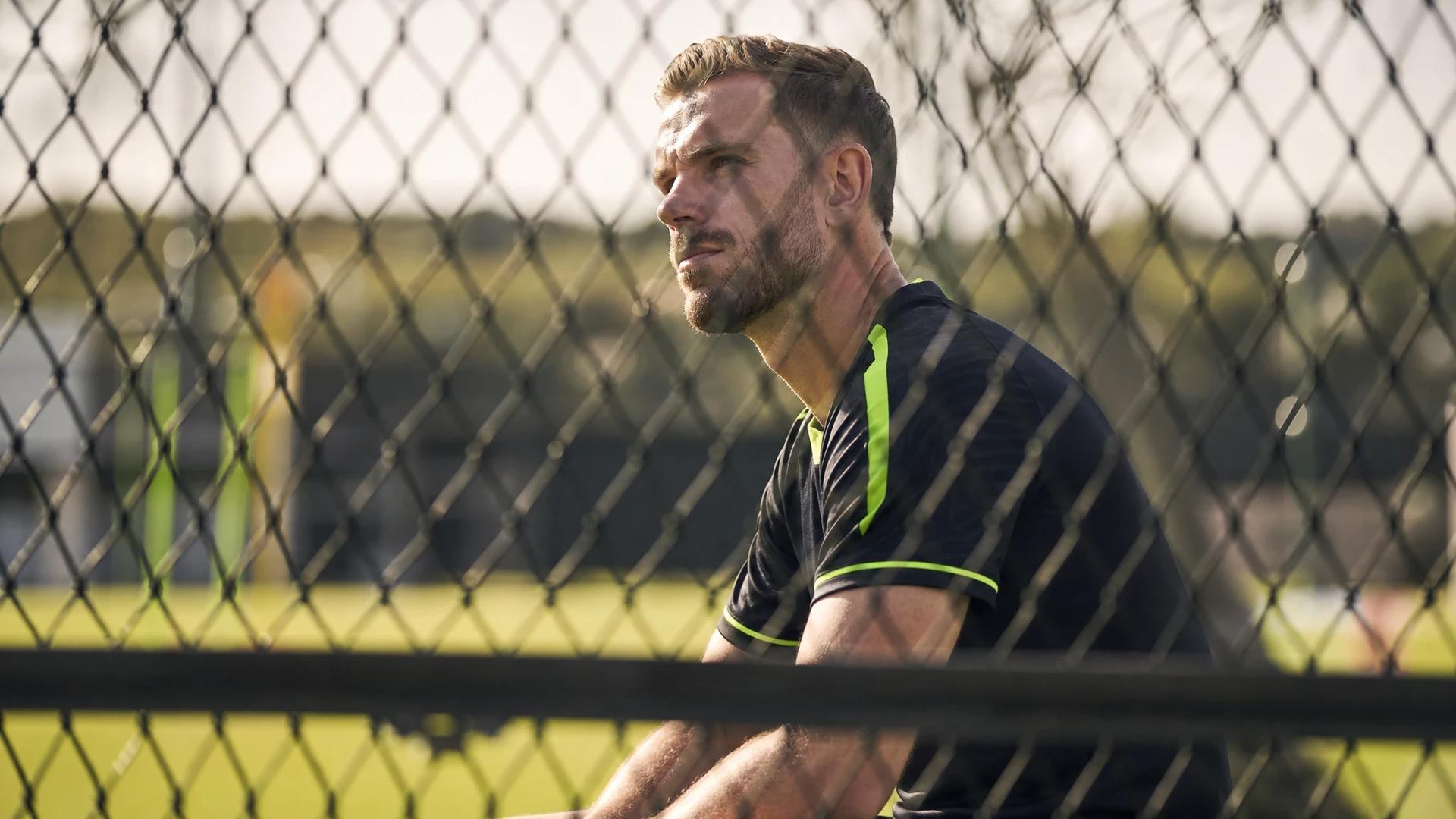 Jordan Henderson standing alone in reflective thought during training, framed through a chain-link fence.