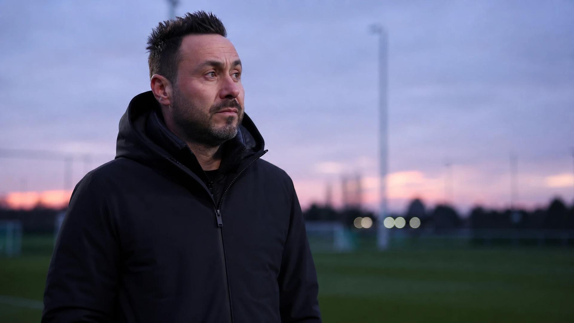 Roberto De Zerbi walking thoughtfully on a training ground during dusk.