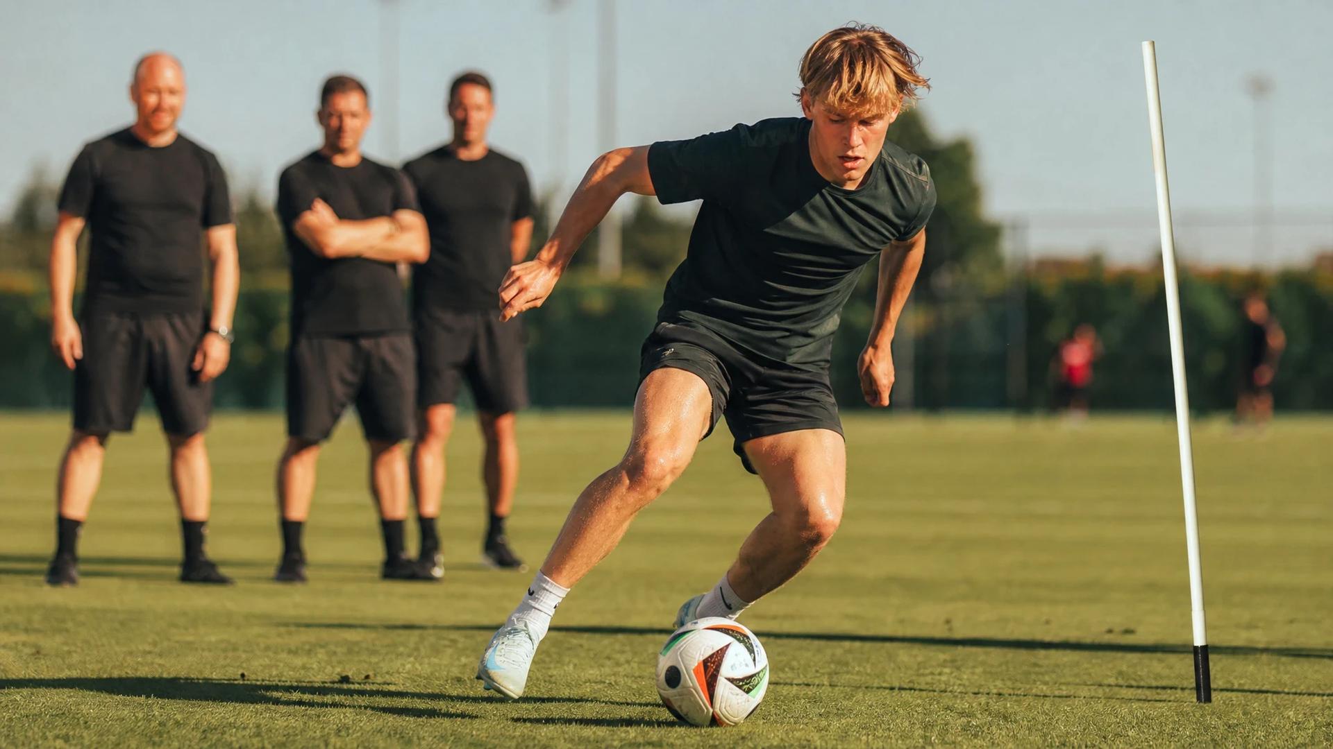 Mika Godts in a training session, dribbling a football under bright sunlight.
