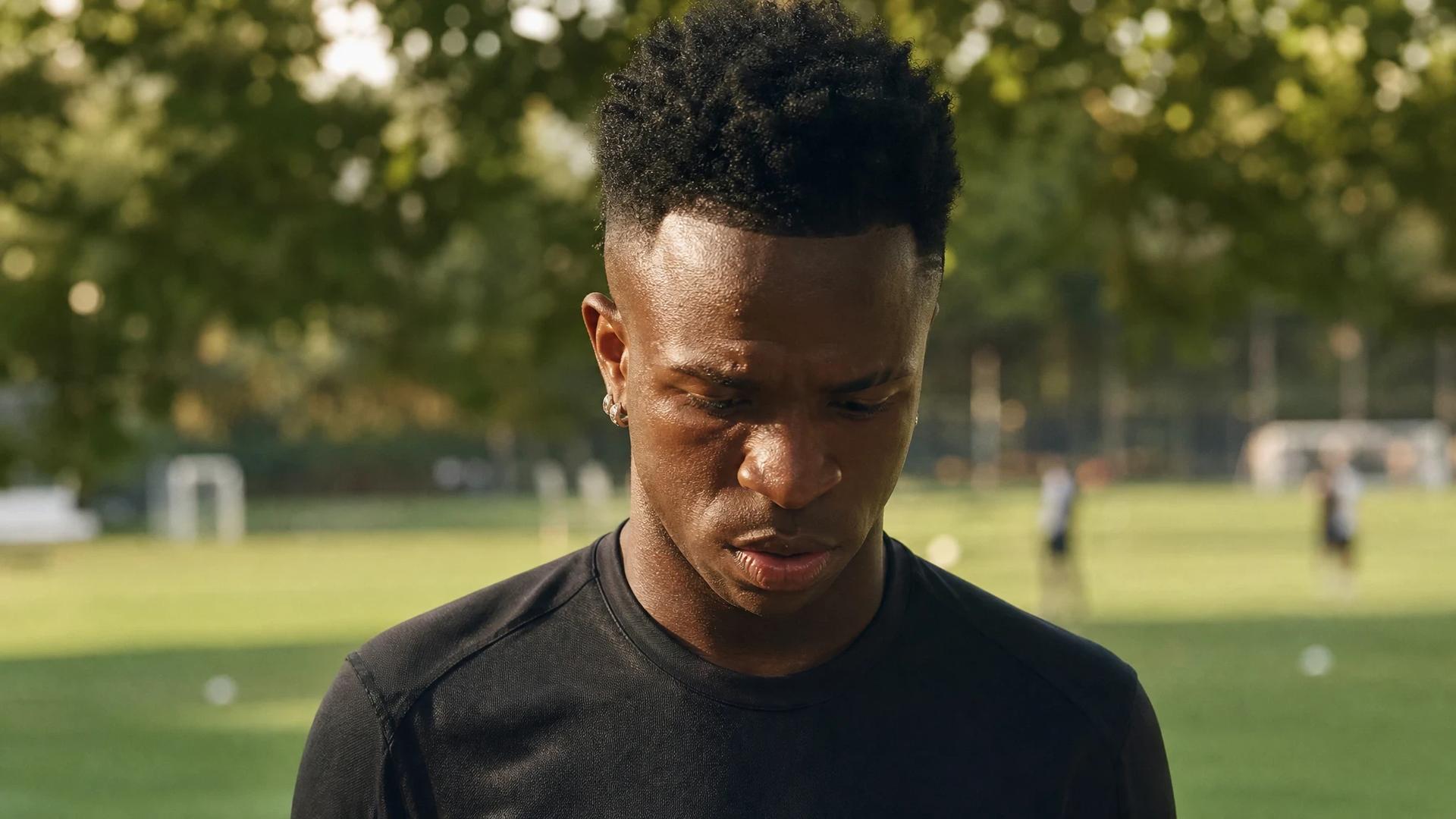 Vinícius Jr. standing thoughtfully in a training ground, surrounded by greenery and soft sunlight.