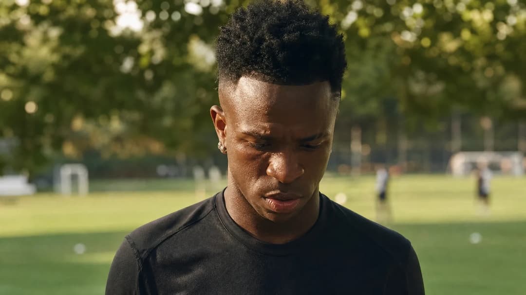 Vinícius Jr. standing thoughtfully in a training ground, surrounded by greenery and soft sunlight.