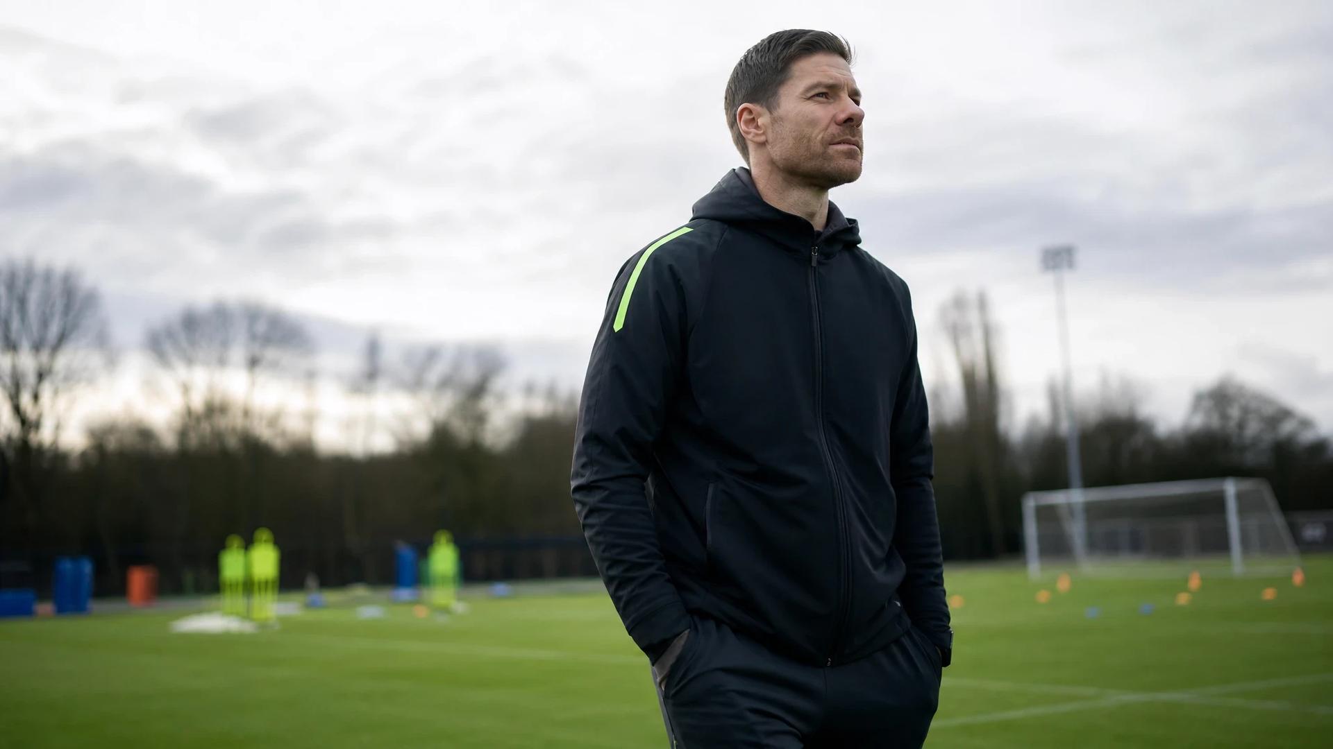 Xabi Alonso stands pensively on the training pitch at Stamford Bridge, surrounded by blurred training equipment.