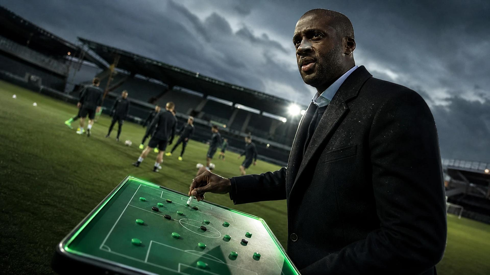 Yaya Touré stands at a tactical board on the sidelines, illuminated by soft artificial lighting.