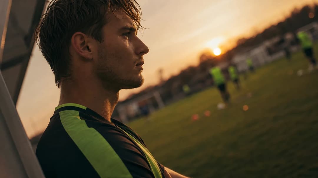A football player stands on a training ground in golden hour light, reflecting on his recent transfer.