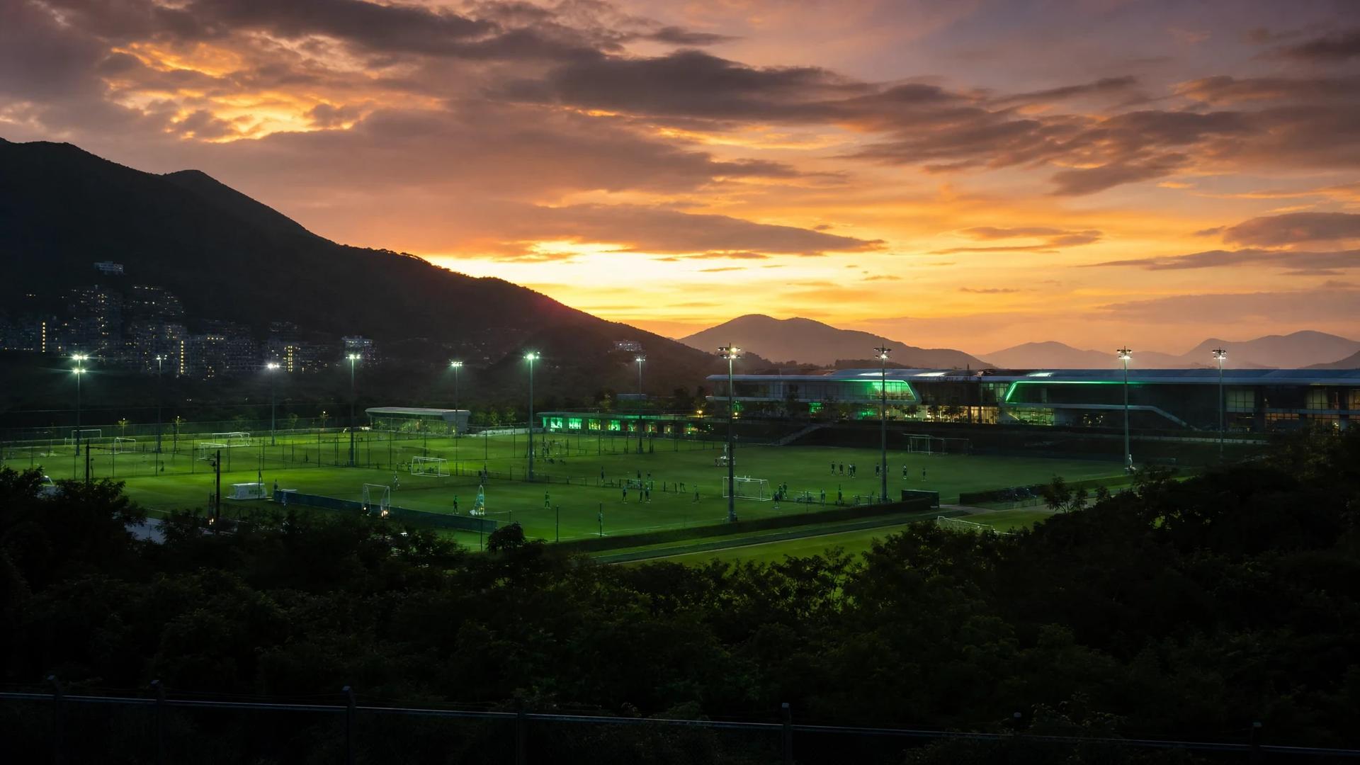 An expansive view of Chelsea's training ground in Hong Kong during the evening, with players silhouetted against neon lights.