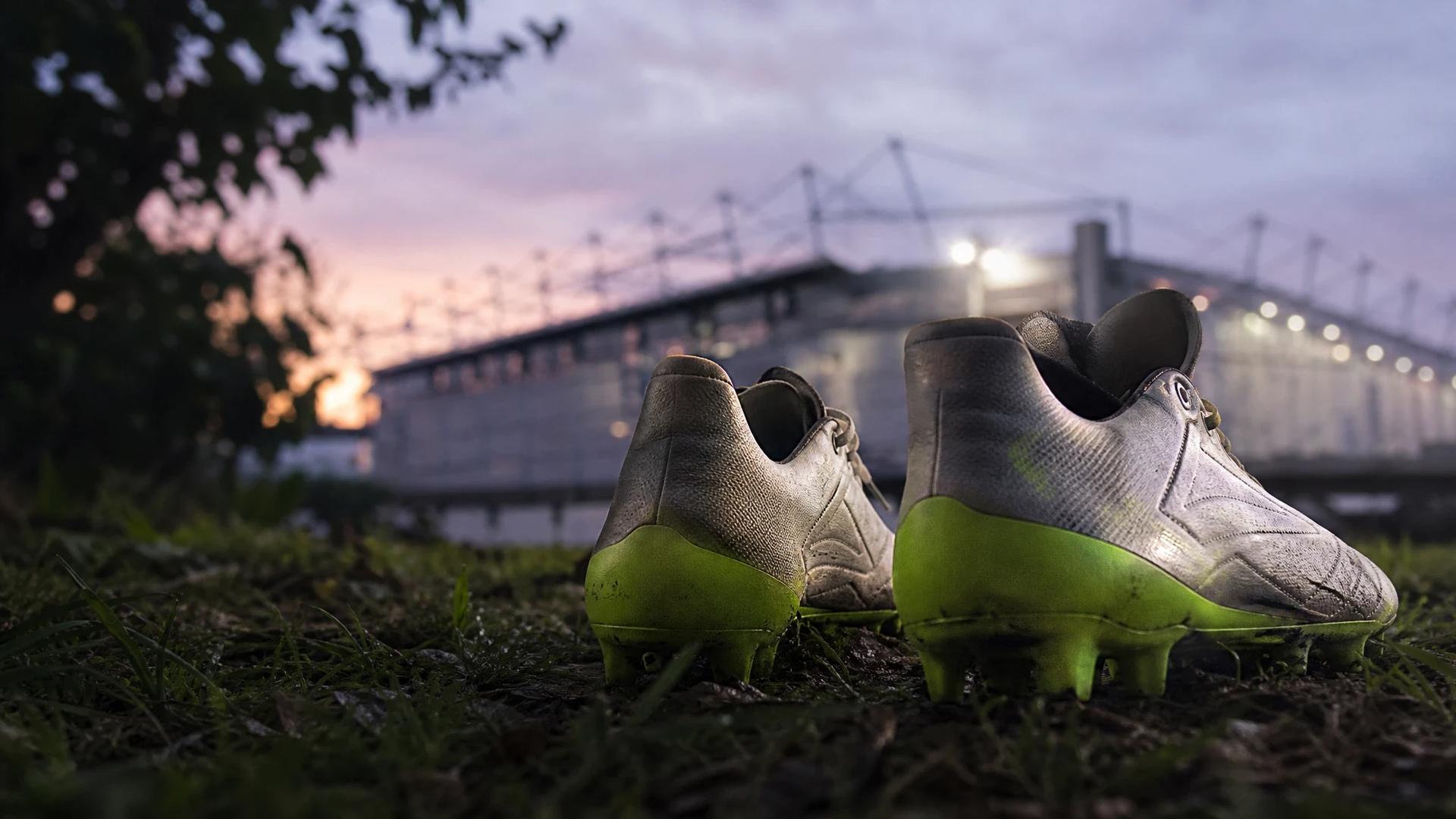 A close-up of football boots resting in front of Goodison Park, illuminated by dappled twilight light.