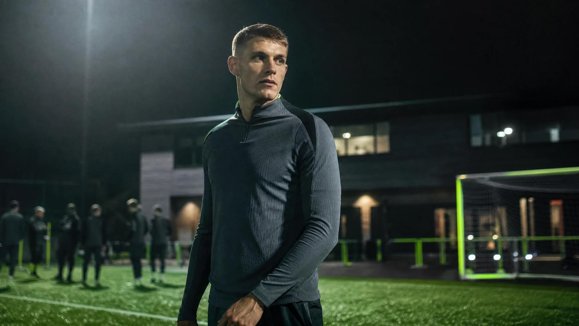 A football player stands on a training pitch with shadows and highlights illuminating his figure.