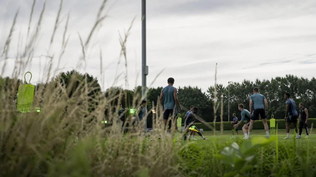 Wide-angle view of Arsenal's training ground with players engaged in practice, framed by tall grass.