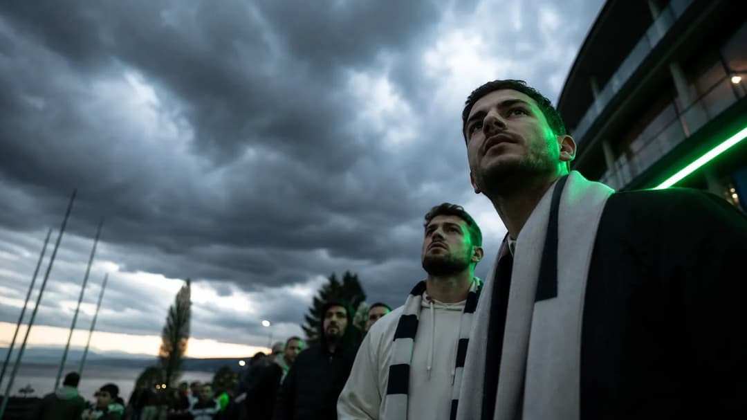 A group of fans in a low-angle shot under stormy clouds, illuminated by neon green accents, eagerly awaiting the Champions League draw.