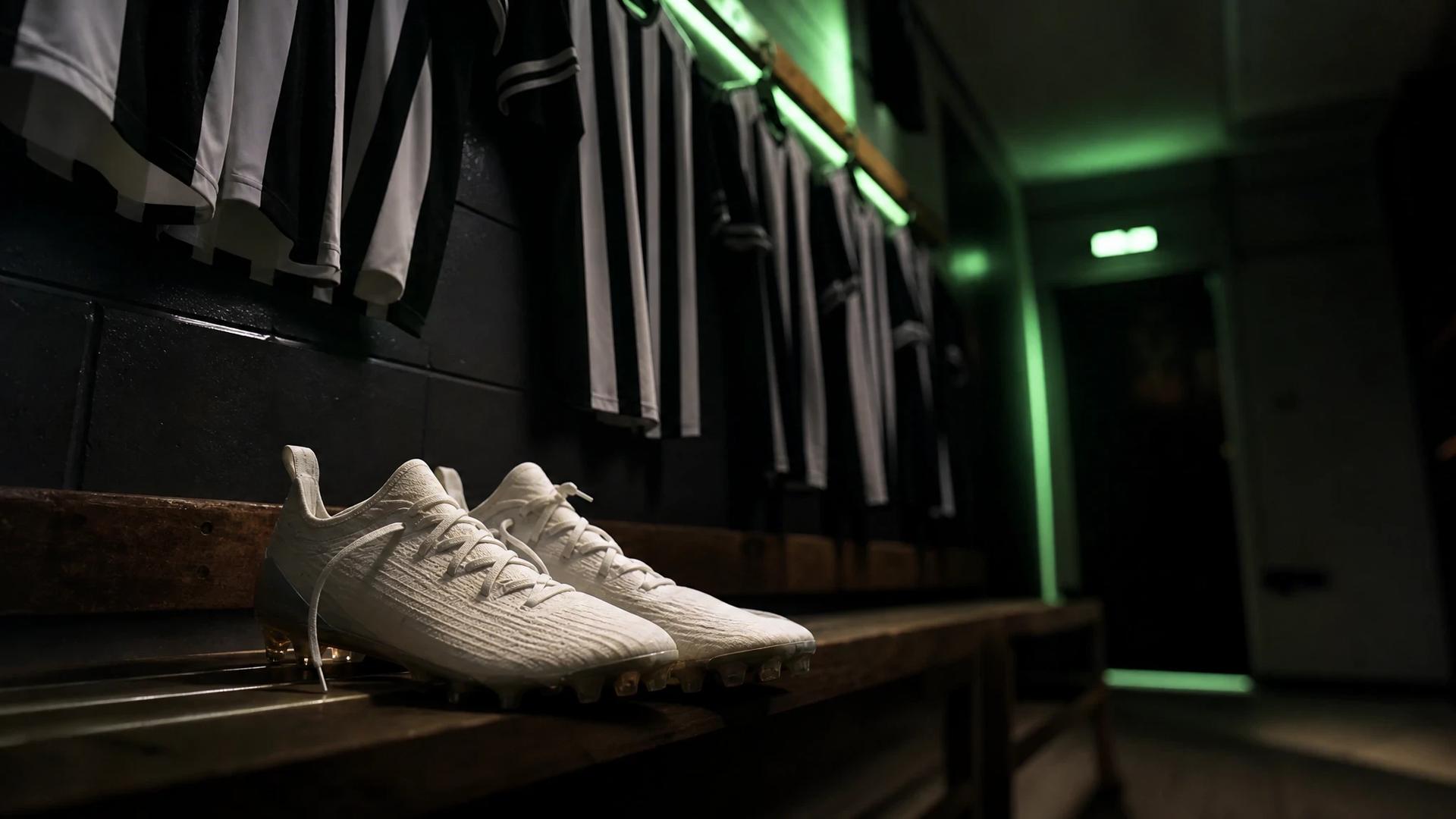 Football boots on a bench in a dimly lit locker room, with jerseys hanging in the background.