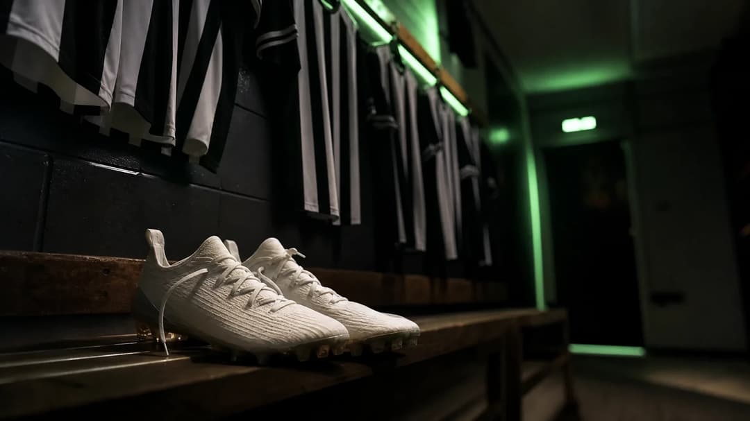 Football boots on a bench in a dimly lit locker room, with jerseys hanging in the background.