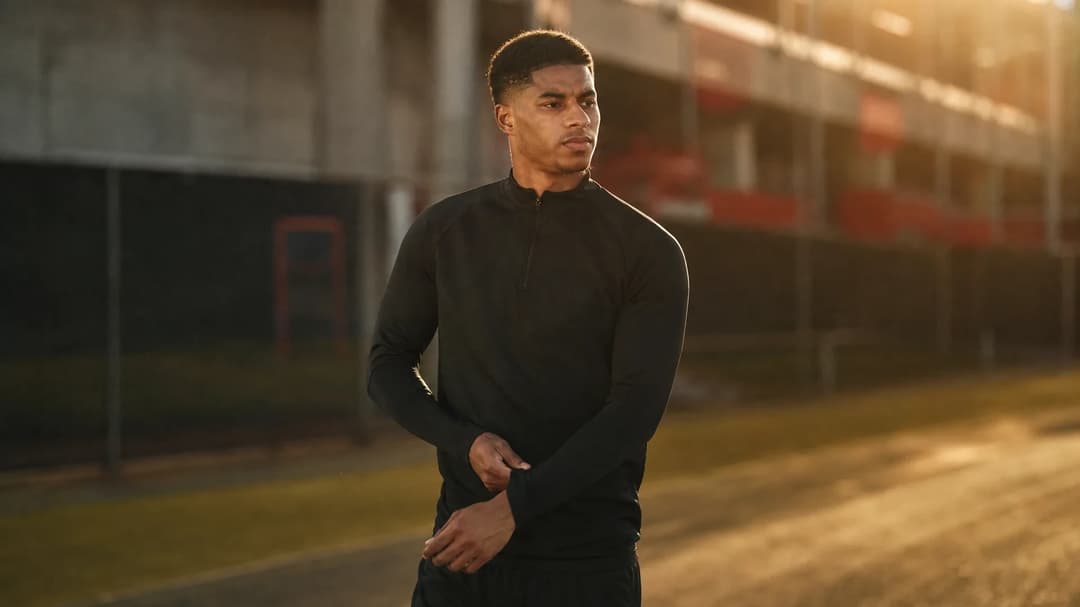 Marcus Rashford stands in a training area, adjusting his gear with a contemplative expression, illuminated by warm lighting.