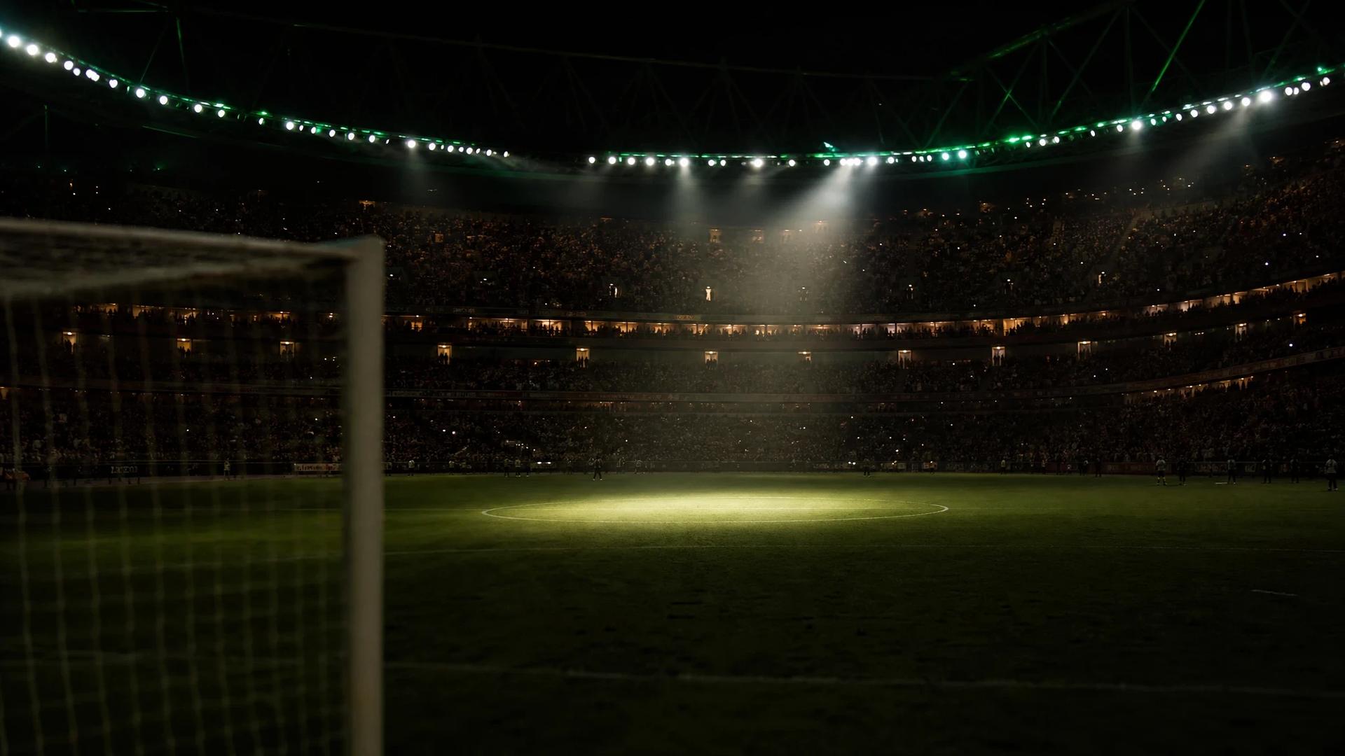 A football stadium scene just before a match, with a goalpost in the foreground and the field illuminated by a single spotlight.