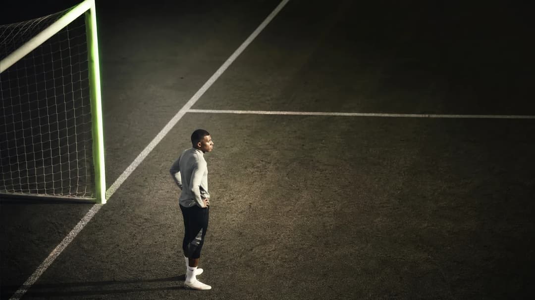 Kylian Mbappé standing contemplatively on a training pitch, viewed from above with soft pastel lighting.