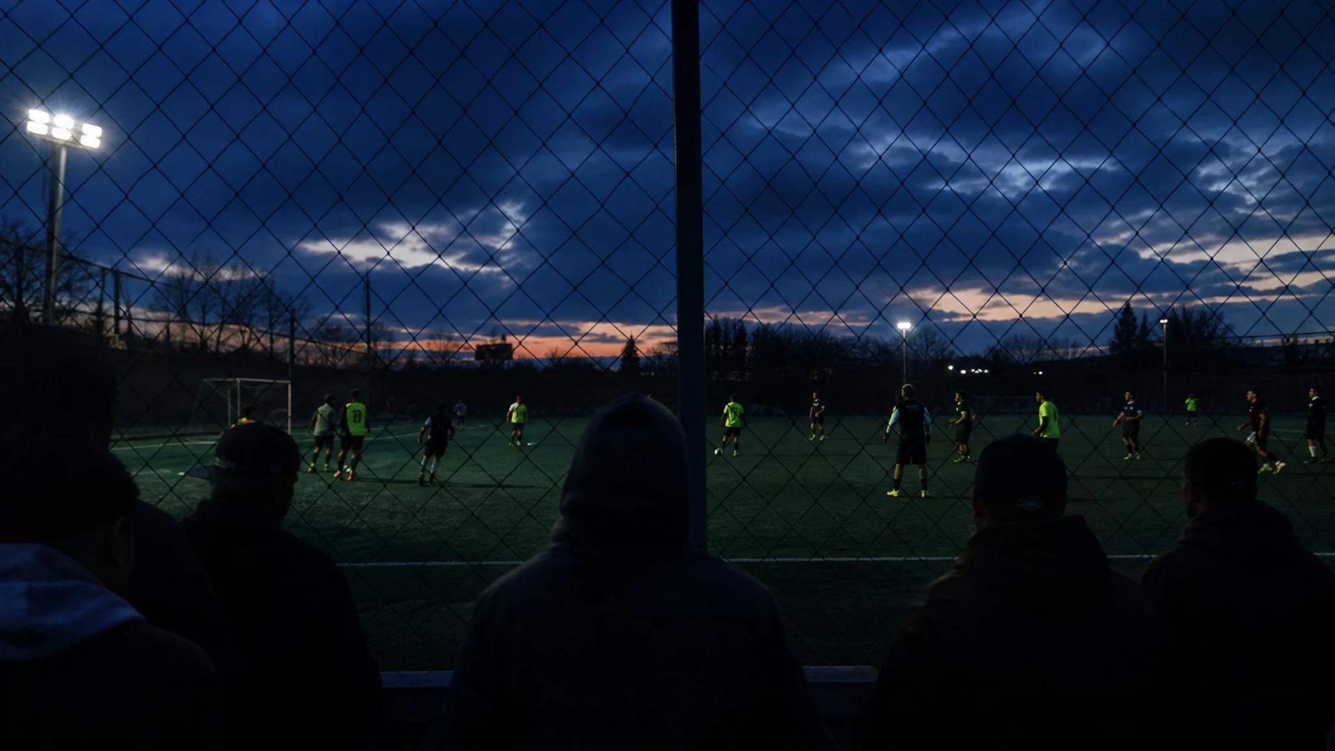 A night scene of Manchester United players training under dim lighting, with shadows of spectators in the foreground.