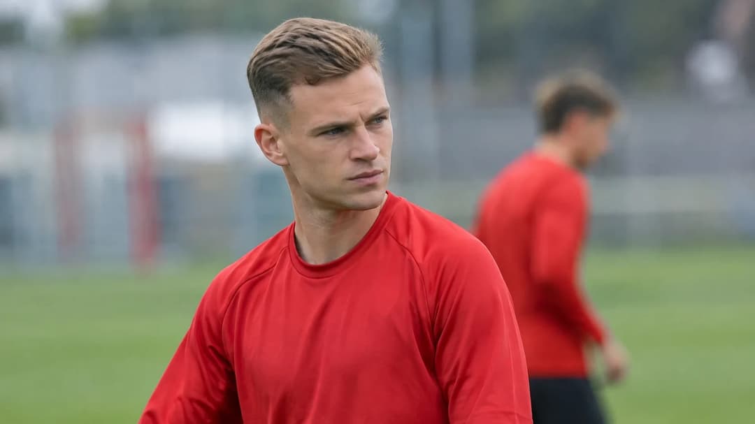 Joshua Kimmich står i et rolig øyeblikk ved en fotballbane, iført en enkel rød treningstrøye.