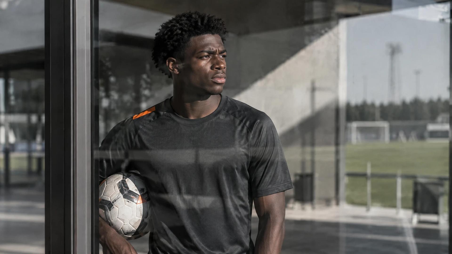 David Otorbi står med en fotball i en solbelyst gang ved treningsbanen.