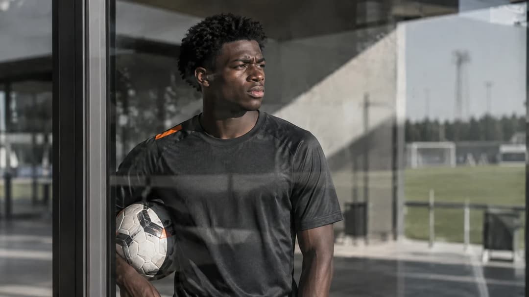 David Otorbi står med en fotball i en solbelyst gang ved treningsbanen.