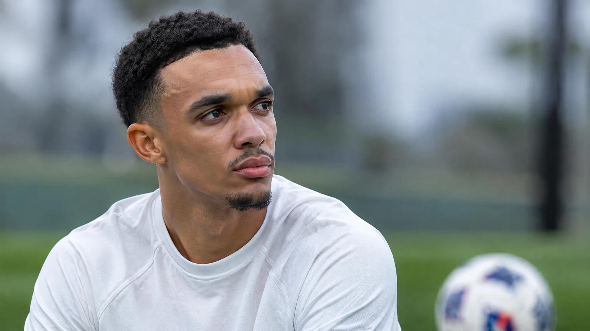 Trent Alexander-Arnold står i en ettertenksom pause ved en fotballbane, med en ball ute av fokus i forgrunnen.