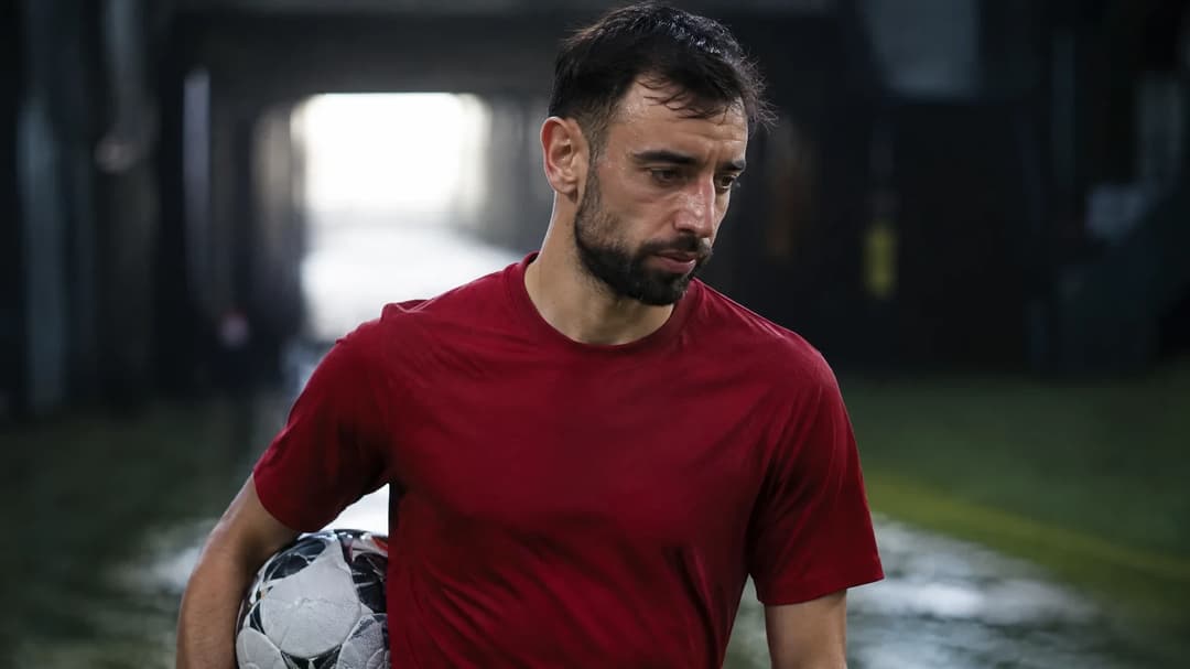 Bruno Fernandes står i et mørkt fotballstadion med en ball under armen og et ettertenksomt uttrykk.