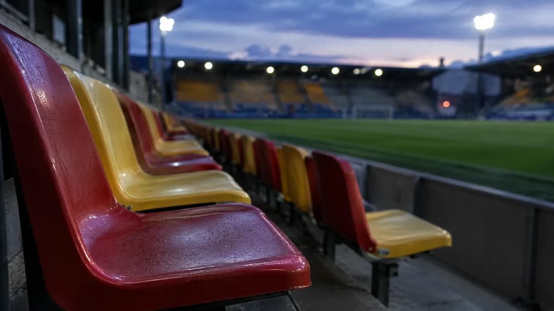 Tomme røde og gule stadionplasser står i fokus foran en uskarp fotballbane i skumringen.