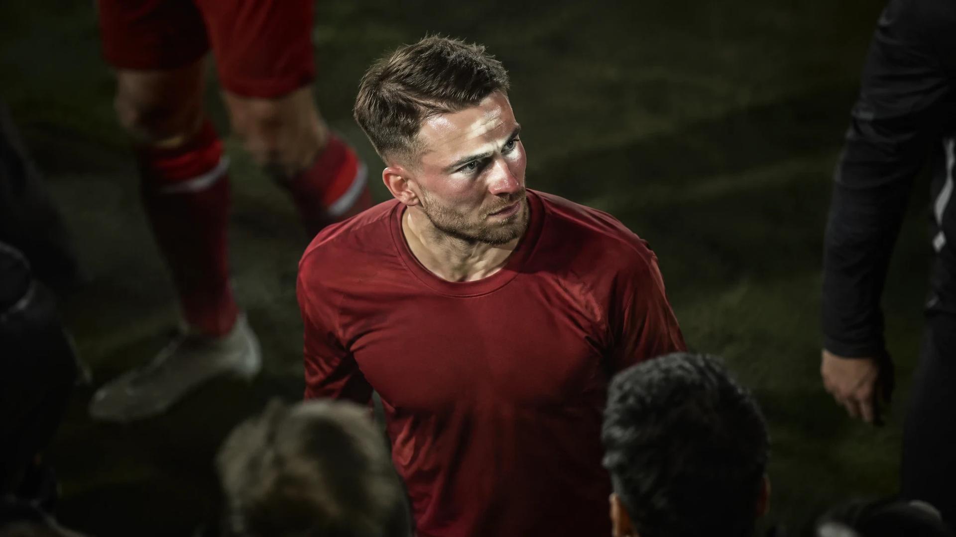 Alexis Mac Allister står på en fotballbane i en plain rød drakt, omgitt av en uskarp folkemengde.
