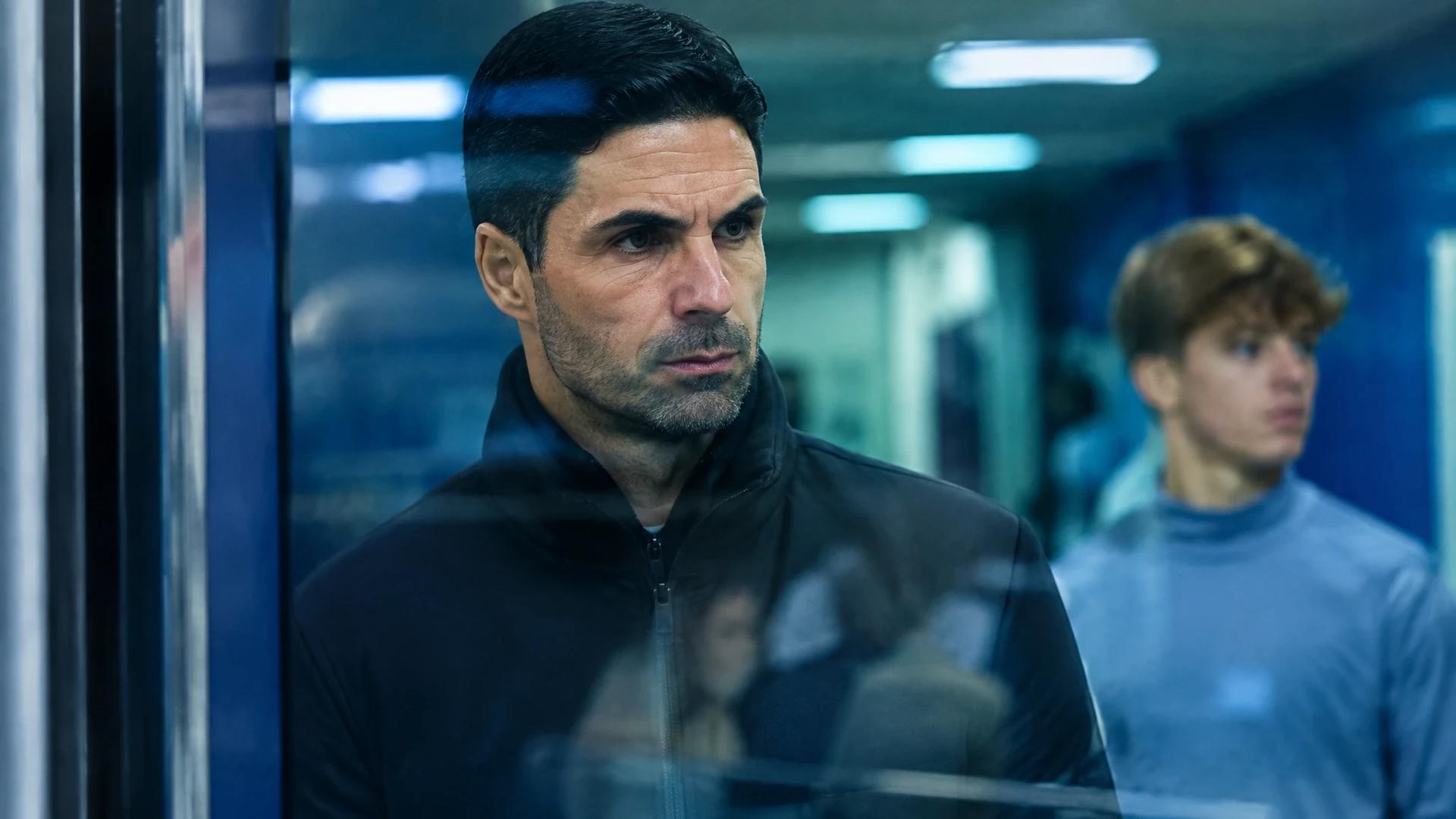 Mikel Arteta står alvorlig i en fotballstadiontunnel, sett gjennom glass, mens en ung spiller er uskarp i bakgrunnen.