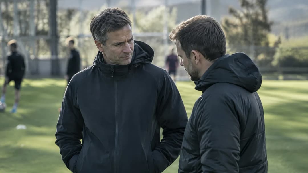 En trener snakker med en medarbeider ved en fotballbane mens spillere er uskarpe i bakgrunnen.