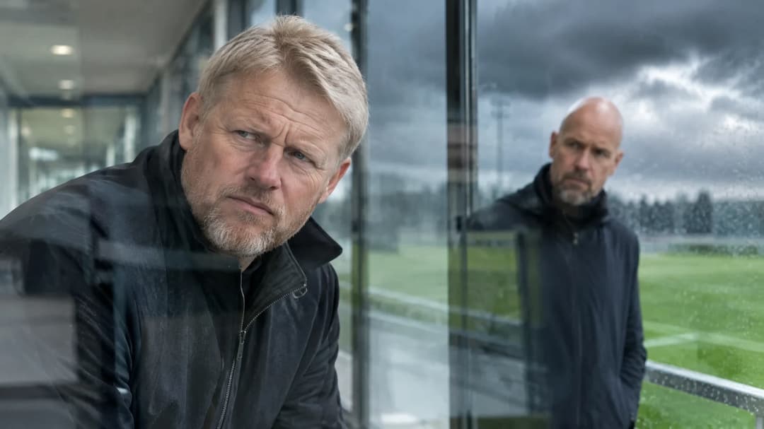 Peter Schmeichel står i forgrunnen i en fotballkorridor, mens Erik ten Hag er uskarpt synlig bak ham gjennom glass.
