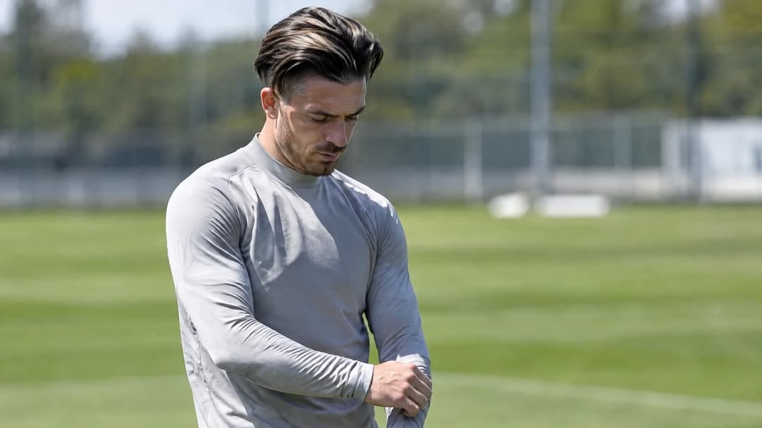 Jack Grealish står alene ved sidelinjen på en treningsbane i sterkt dagslys.