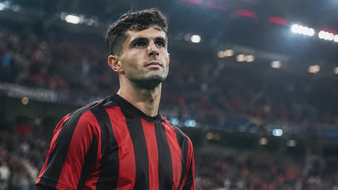 Christian Pulisic står i en umarkert rød-og-svart fotballdrakt på en bane, med et uskarpt publikum i bakgrunnen.
