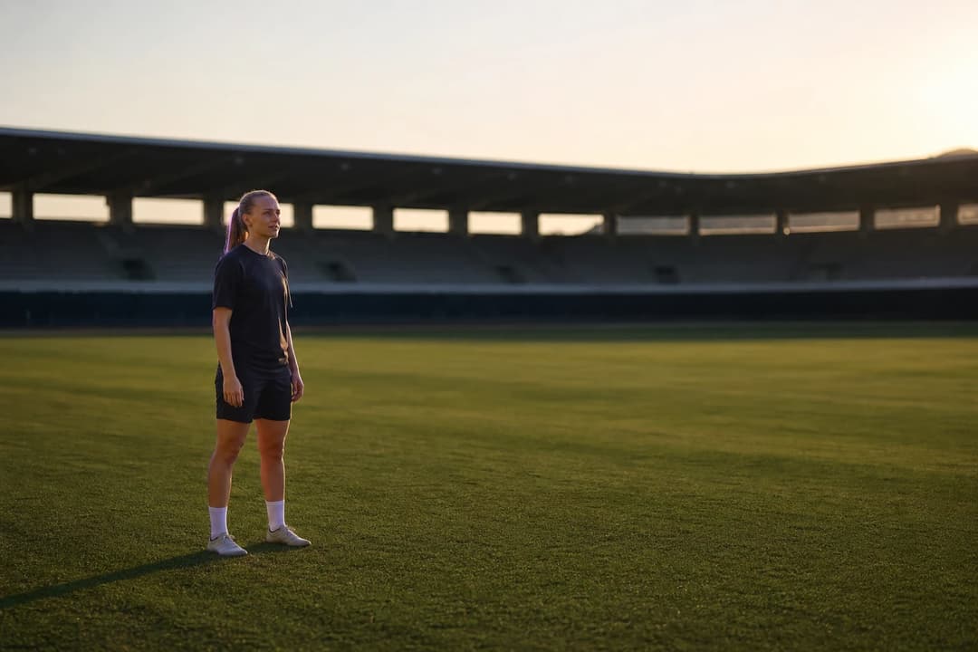 Noelle Maritz stands alone on a training pitch in afternoon light, wearing dark training kit, with a stadium blurred in the distance behind her.