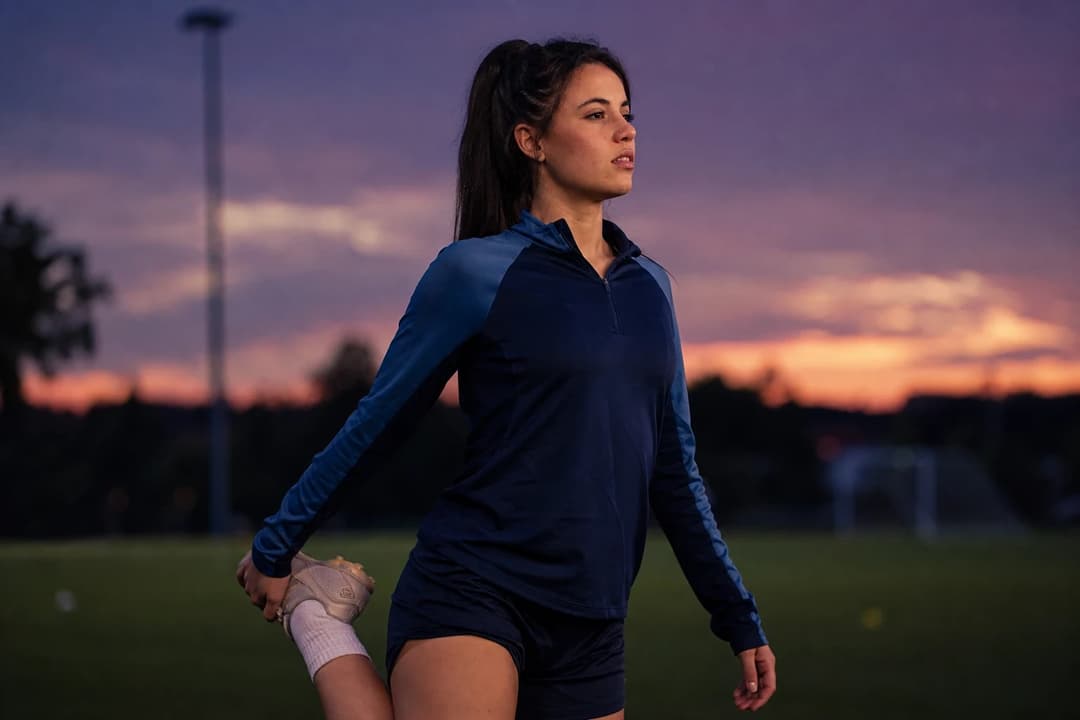 A young female soccer player stretching on a training pitch during twilight.