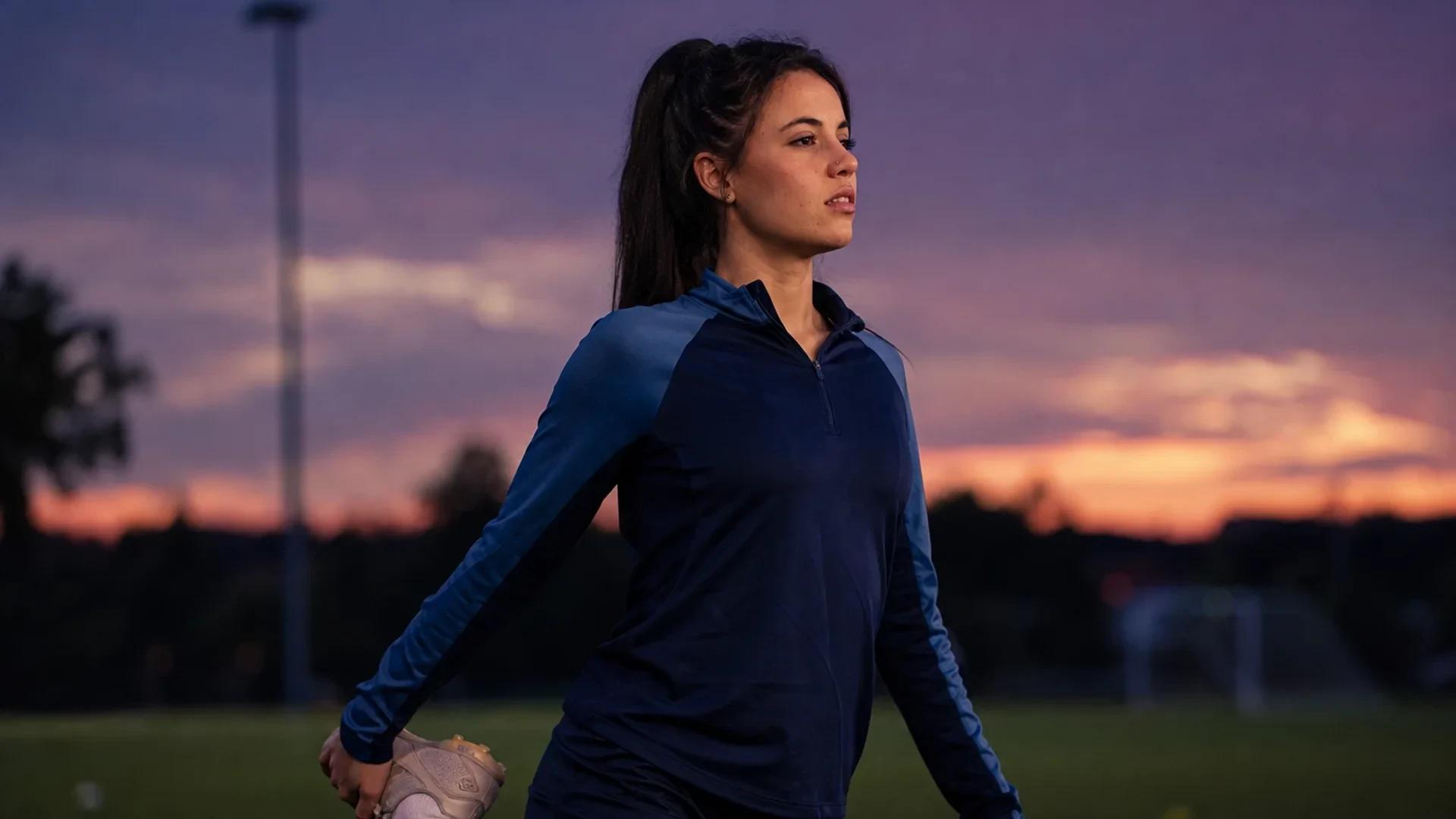 A young female soccer player stretching on a training pitch during twilight.