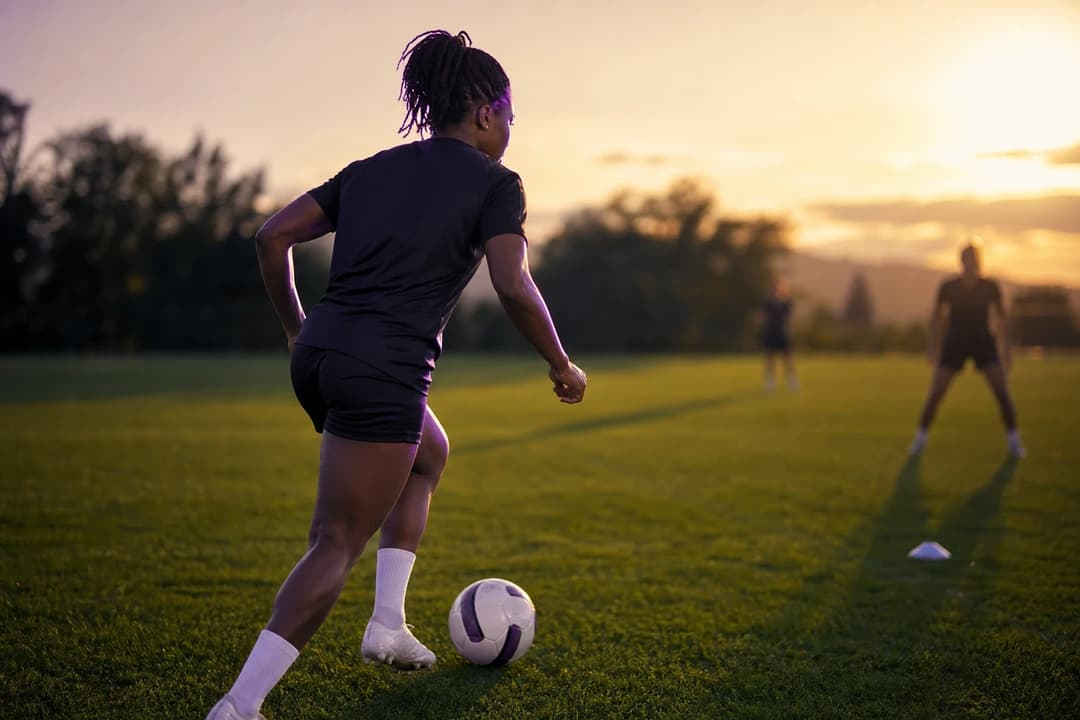 A female footballer practicing dribbling on a training pitch during dusk.