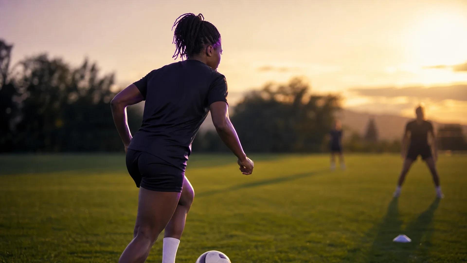 A female footballer practicing dribbling on a training pitch during dusk.