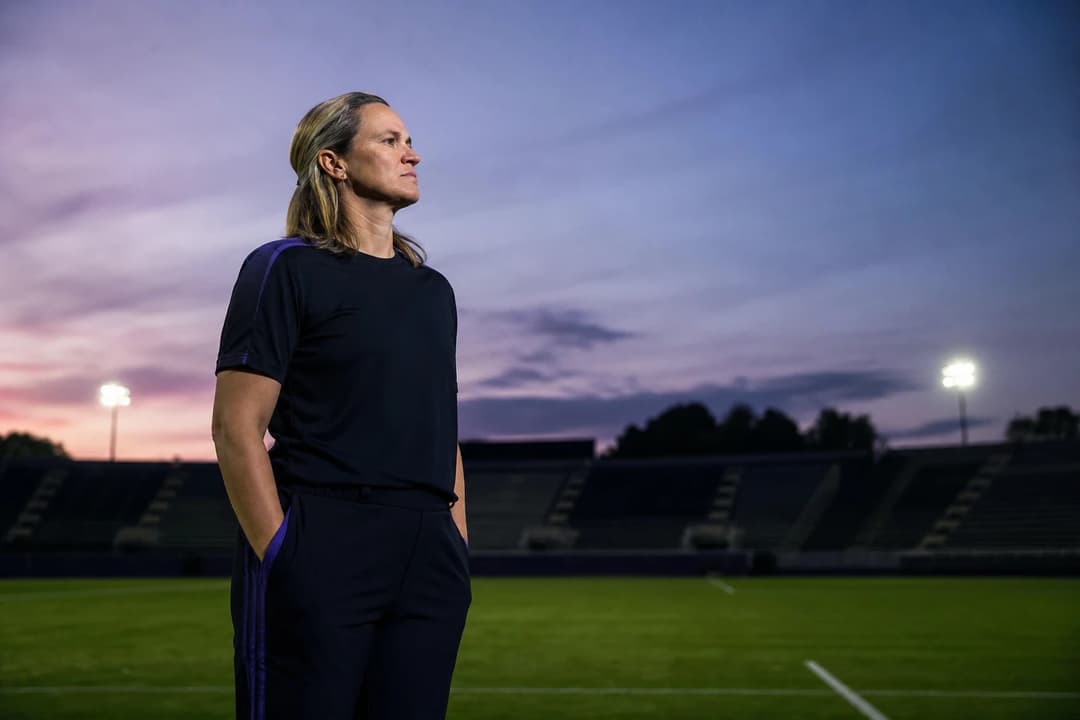 A female soccer player stands alone on the pitch at dusk, contemplating her achievements.