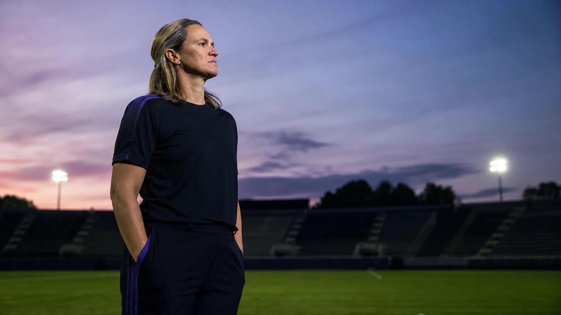 A female soccer player stands alone on the pitch at dusk, contemplating her achievements.