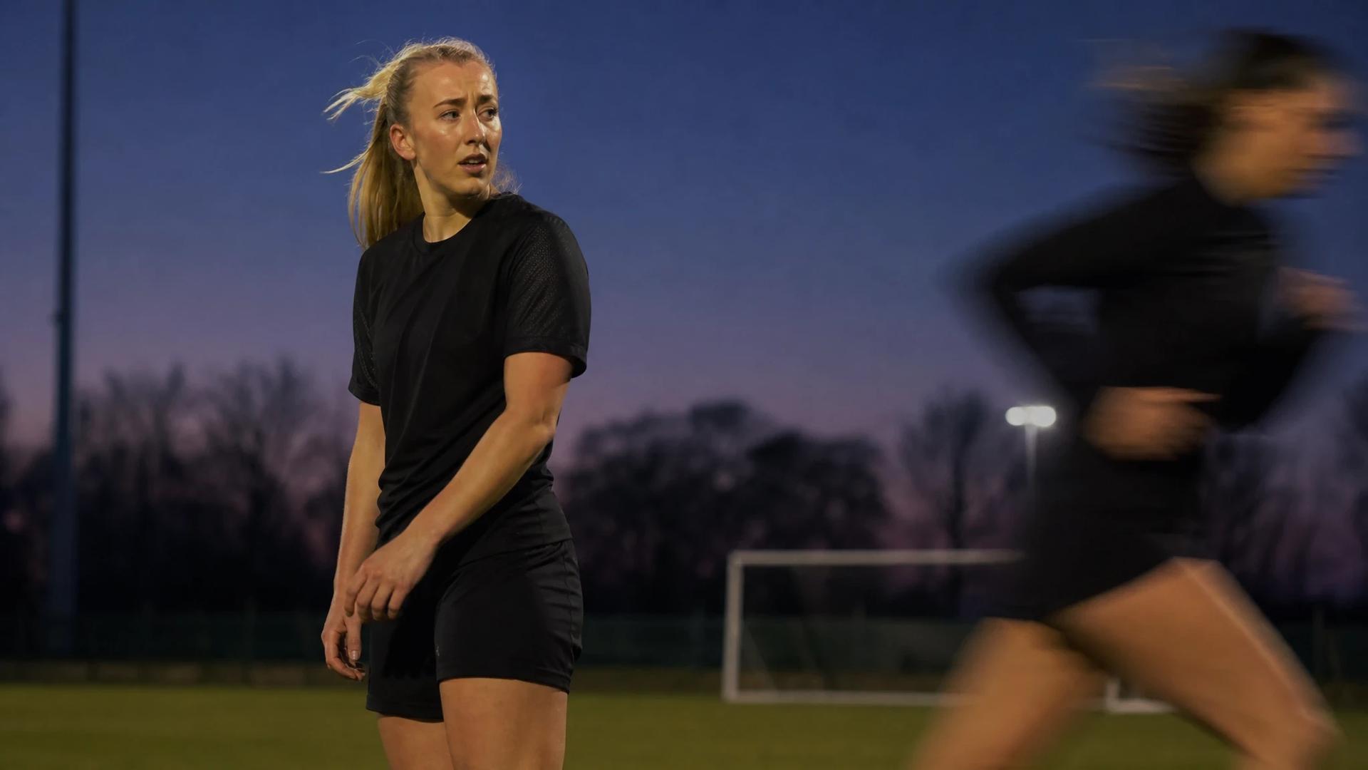 A sportswoman stands poised at dusk on a training pitch, with a teammate blurred in motion behind her.