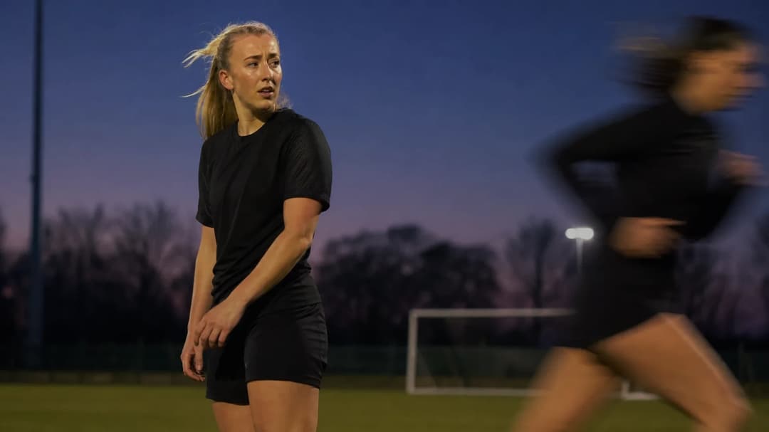 A sportswoman stands poised at dusk on a training pitch, with a teammate blurred in motion behind her.