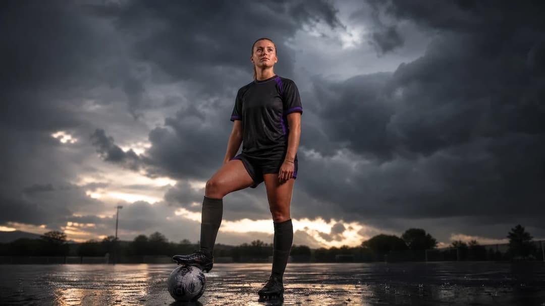 A young female football player looking up on a wet training pitch under stormy clouds.