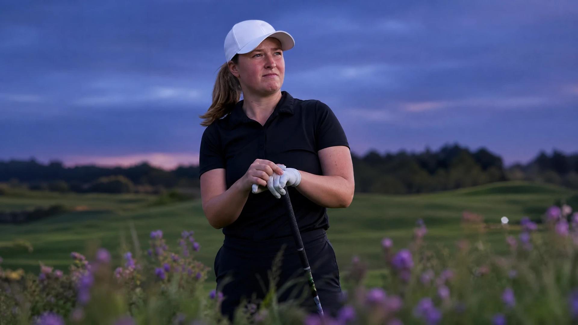 A woman golfer adjusting her club at dusk on a golf course with wildflowers in the foreground.